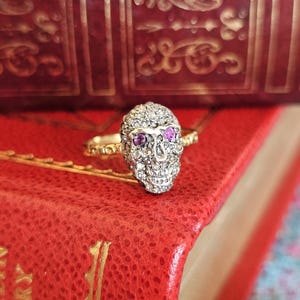 Könnte beinhalten: Ein goldener Ring mit einem totenkopfförmigen Mittelstück, das mit zahlreichen kleinen klaren Steinen und zwei auffälligen rosa Steinen als Augen besetzt ist. Die Ringschiene weist ein texturiertes, perlenartiges Design auf.