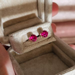 Peut inclure: Une paire de boucles d'oreilles à clous avec des pierres précieuses rouge rubis rondes, serties d'or. Les boucles d'oreilles sont présentées dans un écrin à bijoux beige clair, à la texture douce et veloutée.