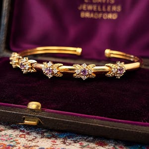 Pode incluir: Uma pulseira de ouro com cinco motivos florais espaçados uniformemente, cada um com uma pedra preciosa redonda roxa central rodeada por pequenas pedras claras. A pulseira é exibida num estojo com o texto E. DAVIS JEWELLERS BRADFORD.