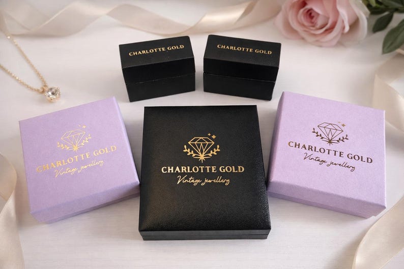 Pode incluir: Uma variedade de caixas de joias em preto e lavanda, com letras e logotipos dourados. As caixas est&atilde;o dispostas sobre uma superf&iacute;cie branca com um colar e uma rosa ao fundo. O texto nas caixas diz "CHARLOTTE GOLD Vintage jewellery."