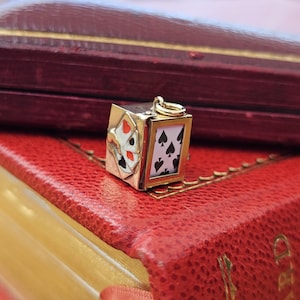 Può includere: Un ciondolo in metallo dorato a forma di piccola scatola, caratterizzato da un pannello bianco con un simbolo di picche nero sul davanti e un pannello laterale decorato con un intarsio in smalto di carte da gioco, inclusi cuori rossi e fiori neri.