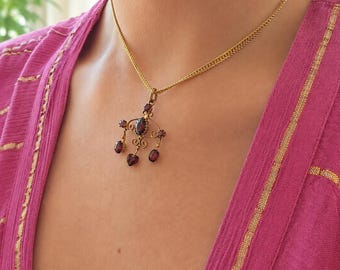 Könnte beinhalten: Eine goldene Halskette mit einer feinen Kette und einem verzierten Anhänger mit fünf dunkelroten Granatsteinen. Der Anhänger hat einen zentralen ovalen Stein, an dem vier kleinere Steine an dekorativen Golddrähten hängen.