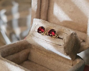 May include: A pair of gold stud earrings featuring round, deep red gemstones. The earrings are presented in a velvet jewellery box with a light beige interior. The box is partially open, showcasing the earrings.