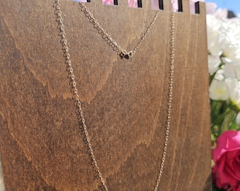 May include: A delicate gold-colored chain necklace displayed on a wooden jewelry stand. The necklace is shown against a backdrop of flowers and a bright blue sky. The necklace is a simple, elegant design.
