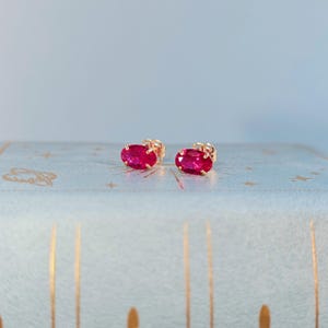 May include: A pair of gold stud earrings, each featuring a vibrant, oval-cut ruby gemstone. The earrings are set against a light blue background with gold star and line accents, creating a delicate and elegant aesthetic.