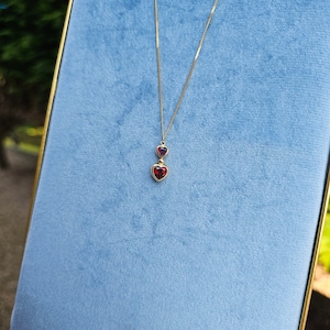 Puede incluir: Un collar dorado con dos colgantes en forma de corazón. El colgante superior es un corazón más pequeño, de color rosa oscuro, y el colgante inferior es un corazón más grande, de color rojo intenso. El collar se muestra sobre un fondo de terciopelo azul claro.