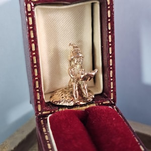 May include: A small gold figurine of a gnome sitting on a mushroom, holding a book. The figurine is in a red velvet box.