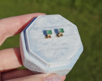 May include: A pair of gold stud earrings with square-cut, vibrant green gemstones, presented in a light blue velvet jewelry box. The earrings are set against a backdrop of green grass.