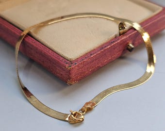 May include: A gold-colored herringbone chain necklace with a clasp. The necklace is displayed on a light gray surface next to a vintage red jewelry box with a beige interior. The necklace is a flat, flexible design.