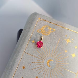 Könnte beinhalten: Ein goldener Anhänger mit einem leuchtend roten Edelstein. Der Anhänger befindet sich vor einem hellblauen Hintergrund mit goldenen Himmelsillustrationen, darunter ein Planet, Sterne und eine Sonne. Das Schmuckstück ist klein und zierlich.