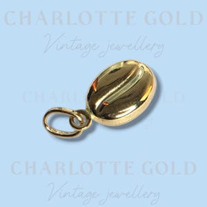 Könnte beinhalten: Ein polierter Goldanhänger in Form einer Kaffeebohne mit einem kleinen befestigten Bindering. Der Hintergrund zeigt den wiederholten Text CHARLOTTE GOLD Vintage jewellery in einem hellblauen Farbschema, passend zum Branding im Bild.