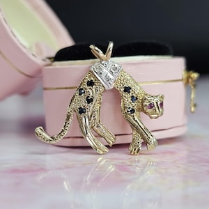 May include: A gold-coloured pendant in the shape of a stylised leopard. The pendant features black and red gemstones, with a diamond-studded section at the centre. The leopard is posed in a crouching position, with its head turned.