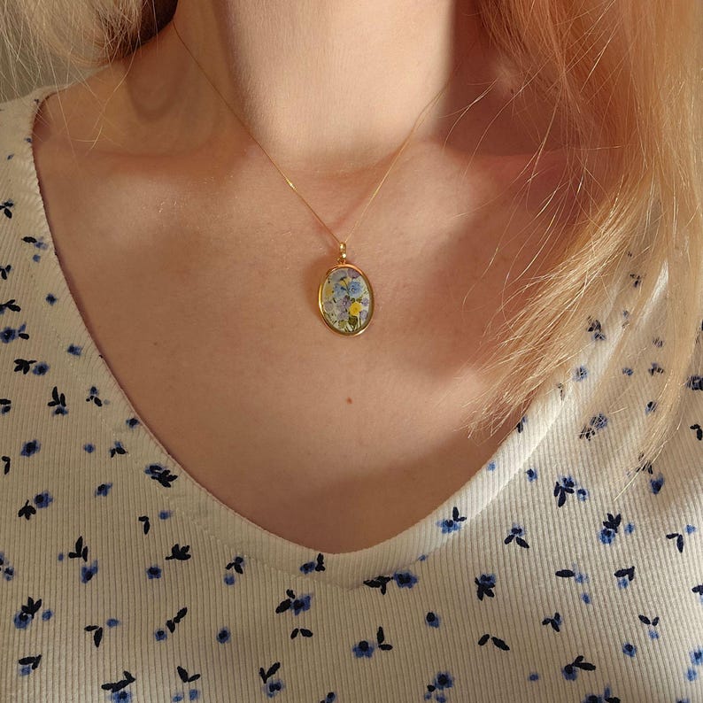 Pode incluir: Um colar dourado com um pingente oval com um design floral. O pingente apresenta pequenas flores coloridas. O colar &eacute; usado sobre uma blusa branca com um padr&atilde;o floral azul.
