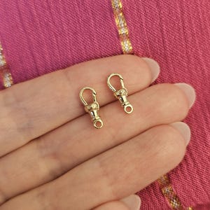 Op de afbeelding: Twee kleine goudkleurige metalen draaibare karabijnsluitingen met een gepolijste afwerking. Elke sluiting is voorzien van een veerbelast mechanisme en een roterende basis, ontworpen voor het maken van sieraden of het bevestigen van accessoires.