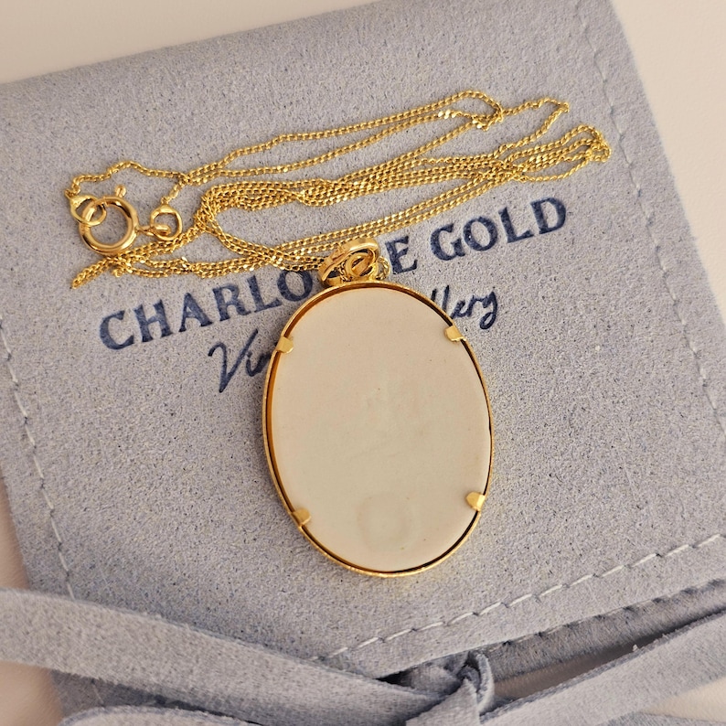 Pode incluir: Um colar dourado com um pingente oval. O pingente tem um centro creme e &eacute; emoldurado por uma borda dourada. O colar est&aacute; em uma bolsa azul clara com o texto "CHARLOTTE GOLD" impresso.