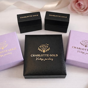 May include: Three square jewelry boxes, two in light purple and one in black, each featuring a gold foil diamond logo and the text Charlotte Gold Vintage jewellery. Two smaller black rectangular boxes are positioned behind the square boxes.