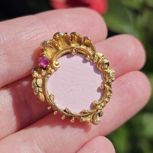 May include: A gold-colored, ornate pendant with a pink center and a small, red gemstone. The pendant has a detailed, floral design around the edges and is held in a hand.