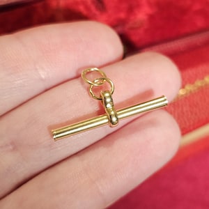 May include: A polished gold-tone metal watch chain T-bar pendant featuring a smooth, cylindrical horizontal bar attached to a small, rounded vertical loop, which connects to two interlocking circular jump rings at the top for secure attachment.