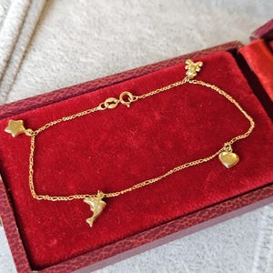 May include: A delicate gold bracelet with charms shaped like a star, dolphin, heart, and a small flower. The bracelet is displayed inside a red velvet jewellery box.
