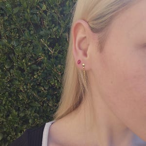 May include: A pair of gold earrings with a small red gemstone on one ear and a longer chain dangling from the other ear.