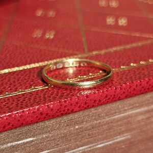 以下が含まれることがあります： 金属の内側に小さな刻印が並んだ、細身で光沢のあるゴールドのバンドリング。リングは滑らかで丸みを帯びた形状をしており、円形の構造全体にわたって均一なイエローゴールドの色調をしています。