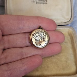 May include: A small, round gold locket with an ornate engraved design. The locket features a detailed floral and shield motif. The locket is held in a hand, and is open, revealing the inside. The locket is in a vintage jewelry box.
