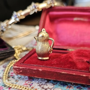 May include: A miniature gold-toned metal charm shaped like a coffee pot or pitcher with a textured, brushed finish. The charm features a rounded body, a small spout, and a curved handle, standing upright as a decorative piece of jewelry.