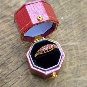 Può includere: Un anello d'oro con pietre preziose rosse, esposto in un portagioie esagonale rosso e oro aperto. L'anello presenta un design ornato con diverse pietre rosse. La scatola è foderata in velluto nero.