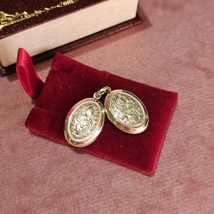 May include: Two gold-colored oval lockets with ornate floral designs rest on a small, red velvet pouch. A maroon book with gold-colored edging is in the background. The lockets are connected by a small loop.