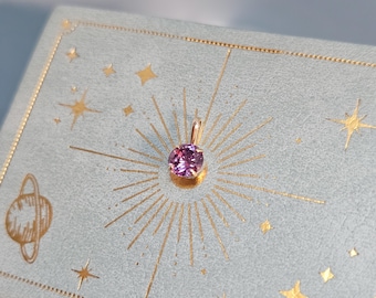 Pode incluir: Um pequeno pingente redondo de pedra preciosa roxa com uma argola dourada. O pingente está sobre um fundo azul claro com desenhos dourados de estrelas e raios de sol. É uma joia.