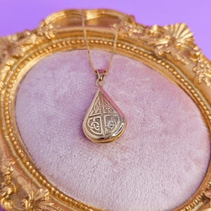 May include: A gold-colored teardrop pendant necklace with an intricate Celtic knot design. The pendant hangs on a delicate gold chain and rests on a pink velvet surface within a gold ornate frame. The background is a solid purple.