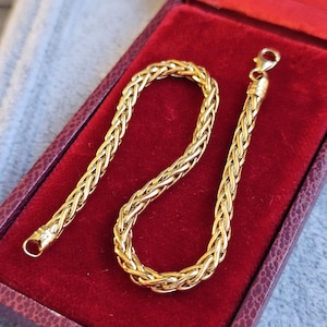 Puede incluir: Una pulsera trenzada de color dorado con cierre de mosquetón, presentada en un joyero de terciopelo rojo. La pulsera tiene un diseño grueso y tejido. La caja está abierta, revelando la pulsera contra el interior rojo.
