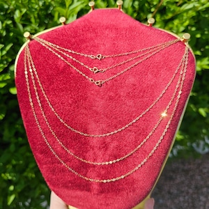 Puede incluir: Varias delicadas cadenas de oro expuestas sobre un expositor de joyería de terciopelo rojo. Las cadenas varían en estilo, algunas con pequeñas cuentas y otras con un diseño de eslabones clásico. Los cierres son visibles, y el expositor está sobre un fondo verde borroso.