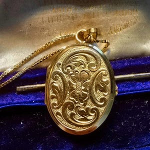 May include: A gold-colored oval locket necklace with an ornate floral design. The locket is suspended from a delicate chain and rests on a dark blue velvet surface. The locket is approximately 1.5 inches in length.