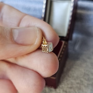 Peut inclure: Un petit charm en forme de dé, de couleur or, avec des détails en émail blanc et des points noirs. Le charm est suspendu à une fine chaîne en or. Le dé est tenu dans une main, avec un écrin bordeaux en arrière-plan.