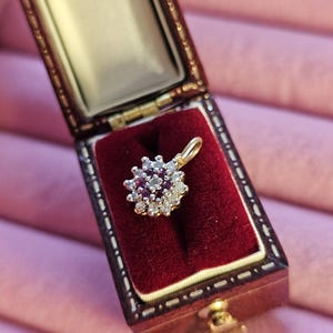 May include: A gold pendant featuring a central cluster of small, red gemstones encircled by smaller, clear stones. The pendant is presented in an open, antique-style jewellery box with a red velvet lining and a brown exterior.