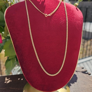 May include: A gold-colored chain necklace displayed on a red velvet bust. The necklace has a delicate curb chain design. The bust is on a gold-colored stand, with pink and white flowers in the background.
