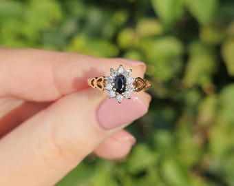 May include: A gold-toned ring featuring a central oval-shaped dark blue gemstone, surrounded by smaller clear stones. The ring has a floral-like design with intricate filigree details on the band. The ring is held against a blurred green background.