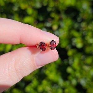 May include: A pair of gold stud earrings, each with an oval-cut, deep red gemstone. The earrings are displayed against a blurred green backdrop, emphasizing the rich colour and sparkle of the stones.