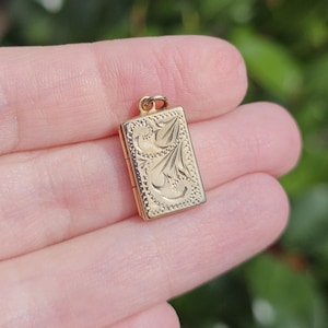 Könnte beinhalten: Ein kleiner, rechteckiger, goldfarbener Anhänger mit einem verzierten floralen Design. Der Anhänger wird zwischen den Fingern gehalten, was seine komplizierten Details und seine filigrane Handwerkskunst zeigt. Der Hintergrund ist unscharf und hebt die Merkmale des Anhängers hervor.