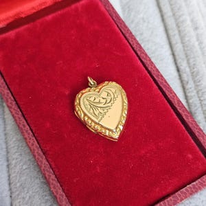 May include: A gold heart-shaped locket with intricate floral detailing, resting in a red velvet lined box. The locket has a decorative border and a small loop for a chain. The box is a deep red color.