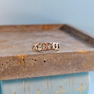 May include: A gold-colored chain link ring. The ring features a series of interconnected links, creating a bold and textured design. The ring is displayed on a concrete surface with a light blue background.