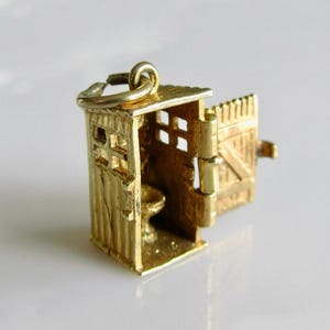 May include: A gold-colored charm in the shape of a small outhouse. The miniature structure features a hinged door, a sloped roof, and a small toilet inside. The charm is designed with detailed vertical siding and small square windows, and includes a loop for attachment.