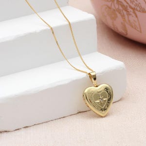 May include: A gold heart-shaped locket pendant suspended from a delicate gold chain. The locket features an intricate engraved scrollwork pattern and a central star motif with a small embedded clear stone at its centre point.