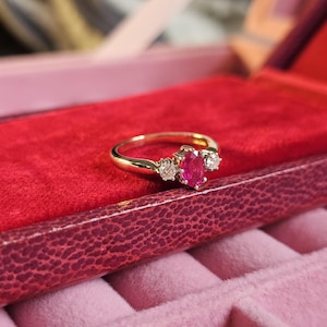 Könnte beinhalten: Ein goldener Ring mit einem ovalen Rubin in der Mitte und zwei kleineren Diamanten auf beiden Seiten, präsentiert in einer roten Samt-Schmuckschatulle. Der Ring hat ein schlankes, poliertes Band. Die Schatulle hat eine strukturierte, dunkelrote Außenseite.