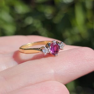 May include: A gold ring with a central oval ruby and two smaller, clear gemstones. The ring has a polished gold band. The gemstones are set in a silver-coloured metal. The background is a blurred green.