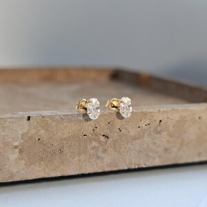 May include: A pair of gold stud earrings featuring oval, clear gemstones. The earrings are displayed on a neutral surface, with a blurred background. These small, delicate earrings are suitable for everyday wear. The gemstones are oval shaped.