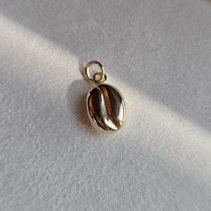 Könnte beinhalten: Ein polierter Goldanhänger in Form einer Kaffeebohne mit einer glatten, reflektierenden Oberfläche, einer zentralen vertikalen Rille und einem kleinen runden Biegering an der Oberseite zum Befestigen an einer Halskette oder einem Armband.