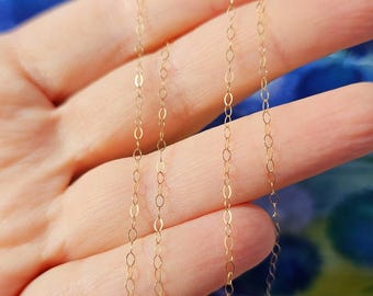 May include: Delicate gold chain necklace with small, oval-shaped links. The necklace is held against a blurred, colorful background. The chain has a lobster clasp closure. A simple and elegant piece of jewelry.