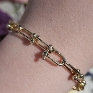 May include: A gold bracelet with a chain-link design. The bracelet features elongated links connected by small, spherical accents. The links are smooth and polished, reflecting light. The bracelet is a piece of jewellery.